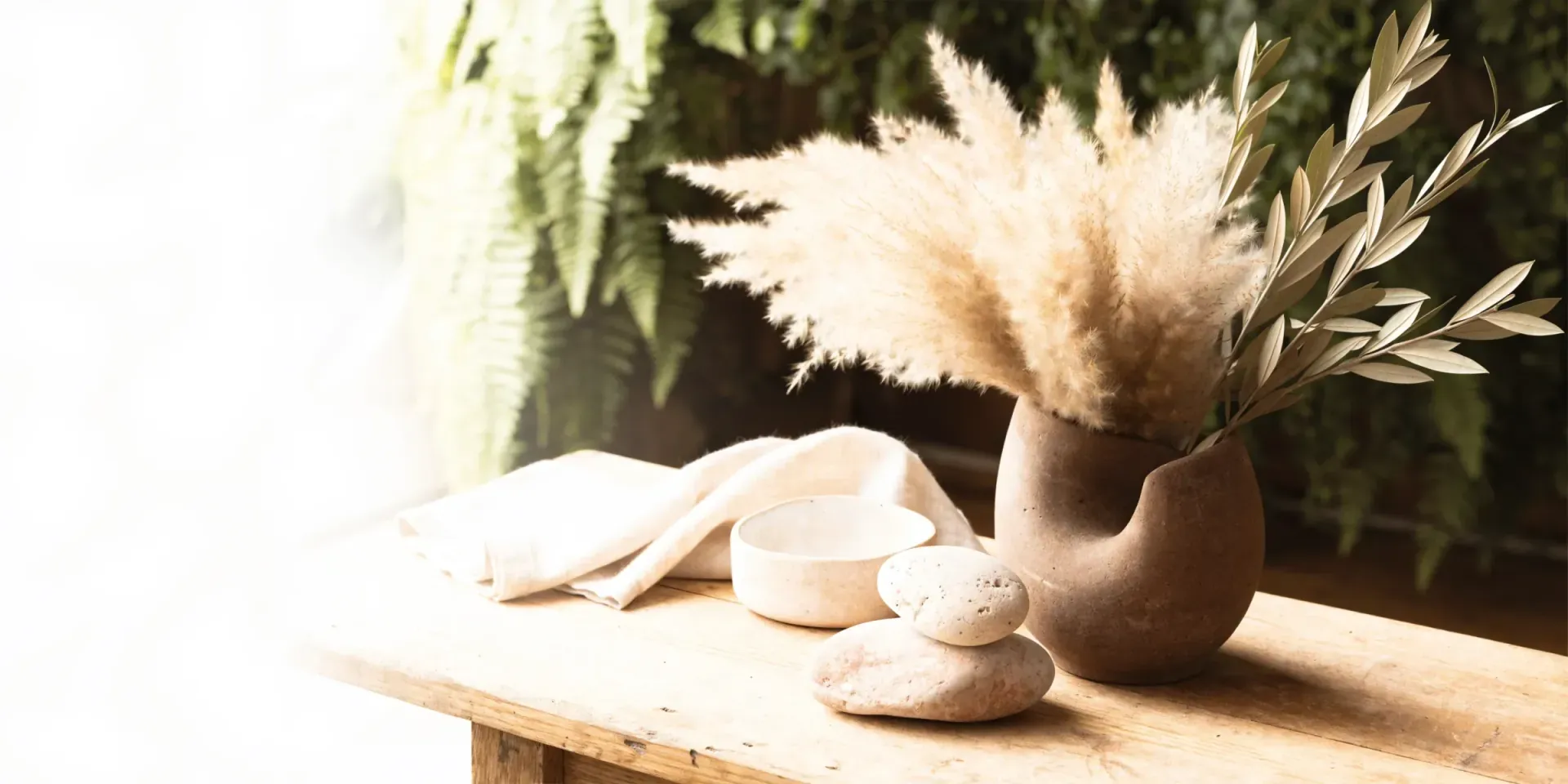 A worn light-oak surface in warm light with folded linen, dried pampas and olive branches in a stoneware vessel, and smooth travertine stones