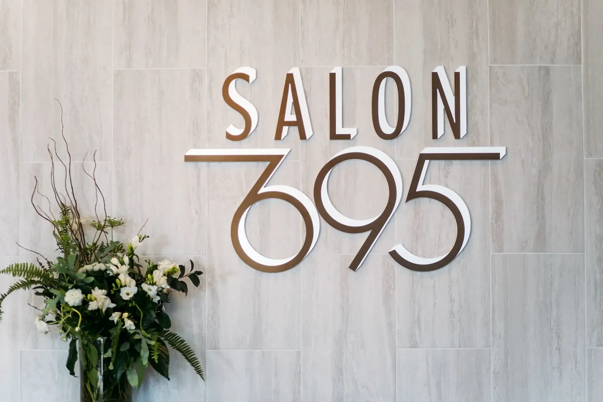 The brushed-metal Salon 695 sign mounted on a pale travertine wall, with a full white-and-green floral arrangement to the left