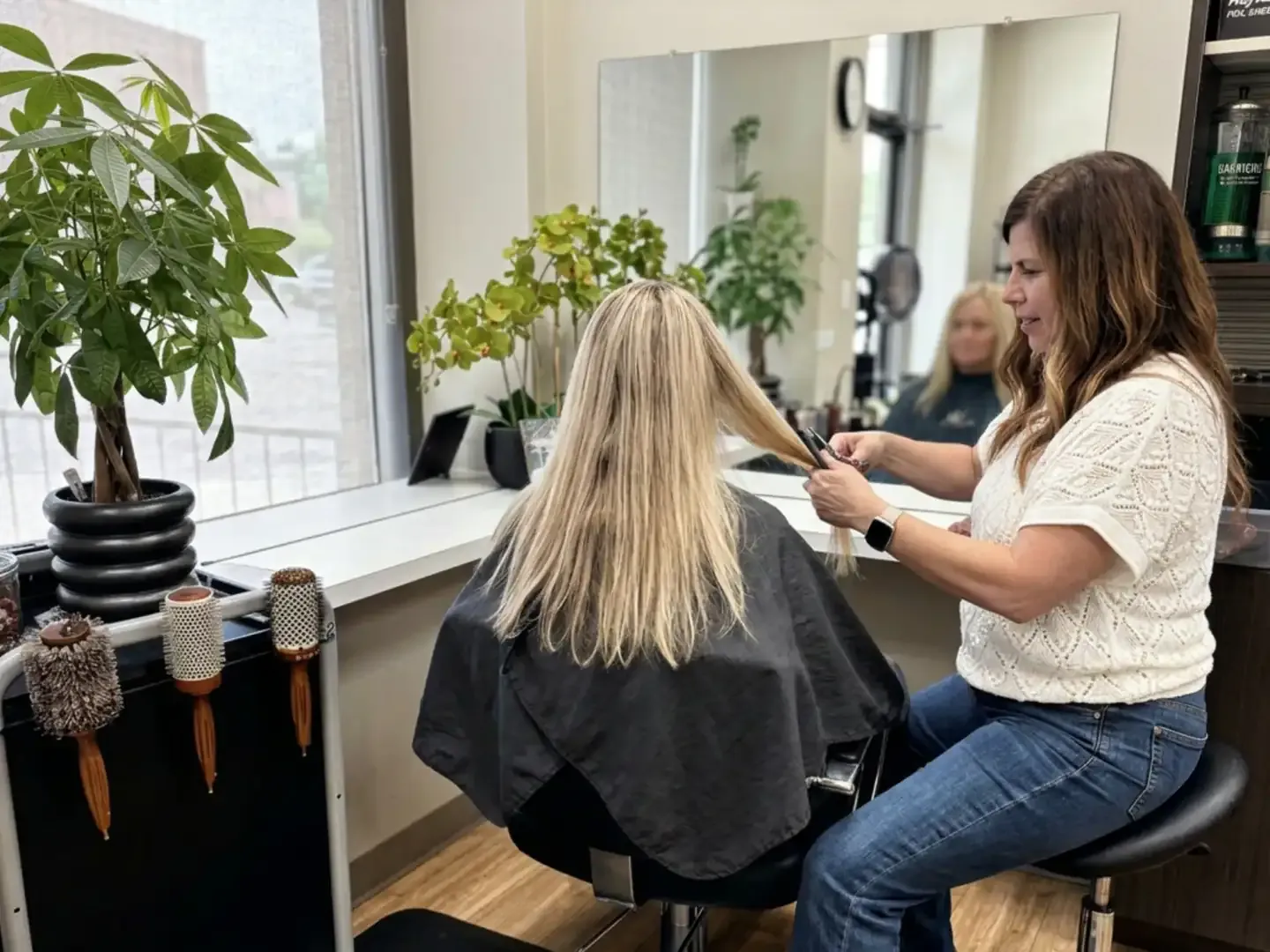 A stylist at her own station cutting a blonde client's long hair by a plant-filled window, styling brushes on the cart and product shelves behind her