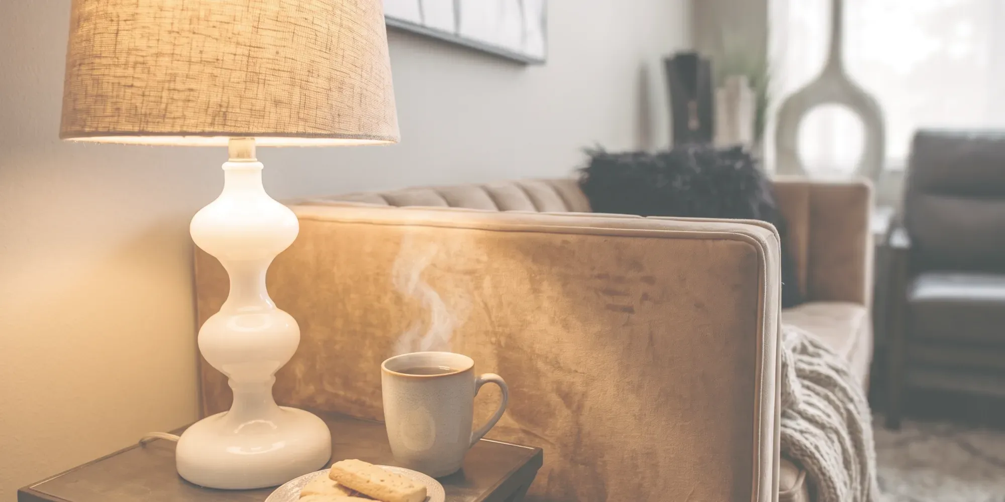 A quiet salon lounge corner under a soft milky light, a lit lamp on a side table beside a steaming mug and shortbread with a tan velvet sofa and a knit throw