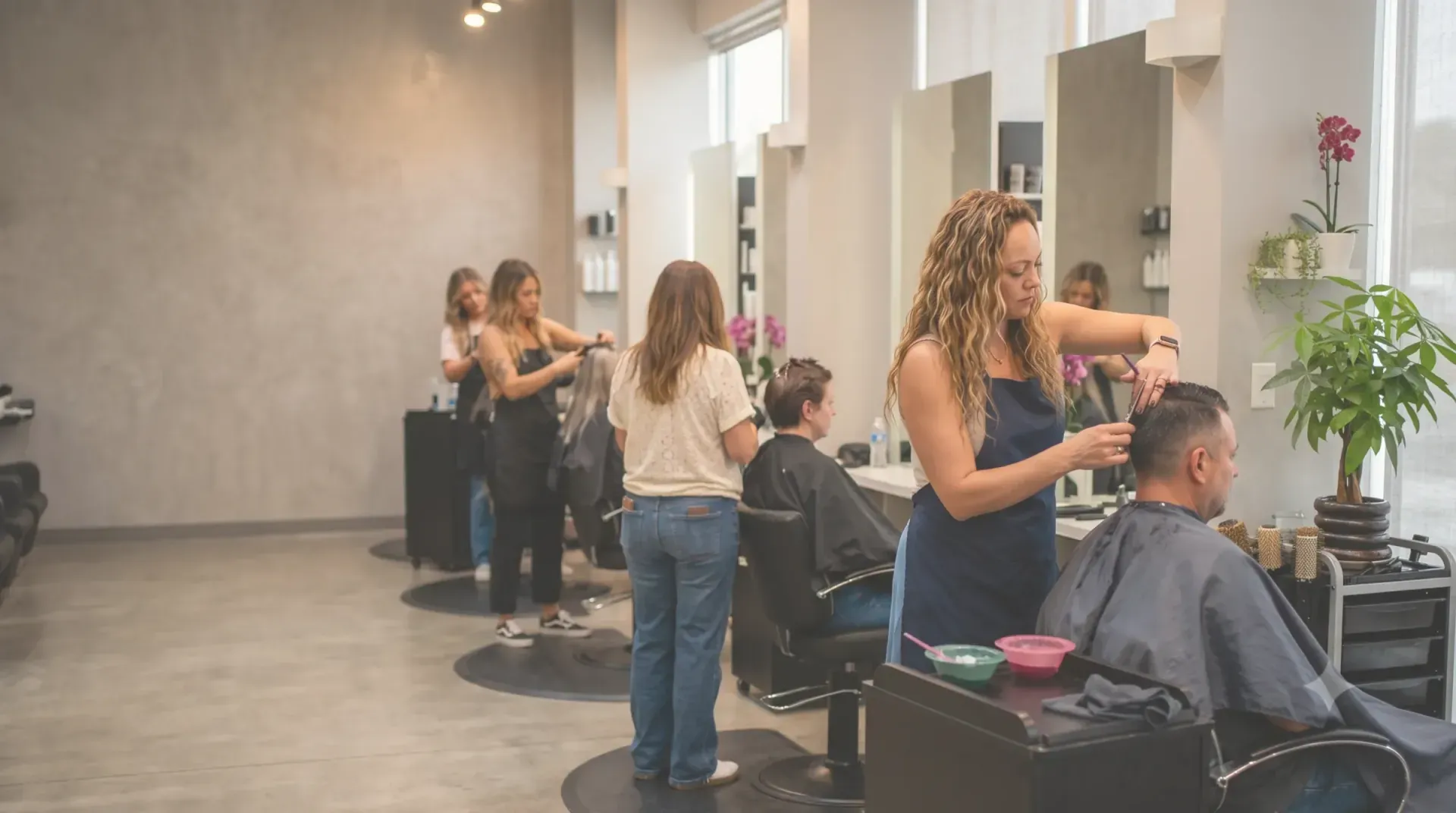 The Salon 695 floor in use: several stylists working at their own stations along a bright wall of mirrors, one cutting a client's hair in the foreground, plants and product shelves throughout the room