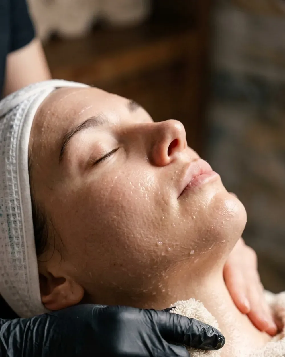 A client with a headband resting during a facial, an esthetician steadying her neck in a black glove