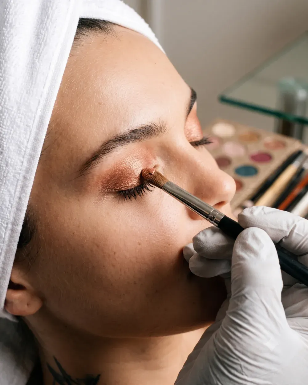 A makeup artist blending warm shimmer eyeshadow onto a client’s lid with a brush, a palette behind