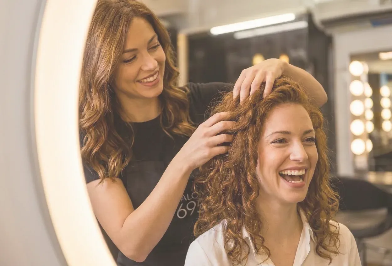 A Salon 695 stylist running her fingers through a laughing client's curly auburn hair under a lit ring mirror