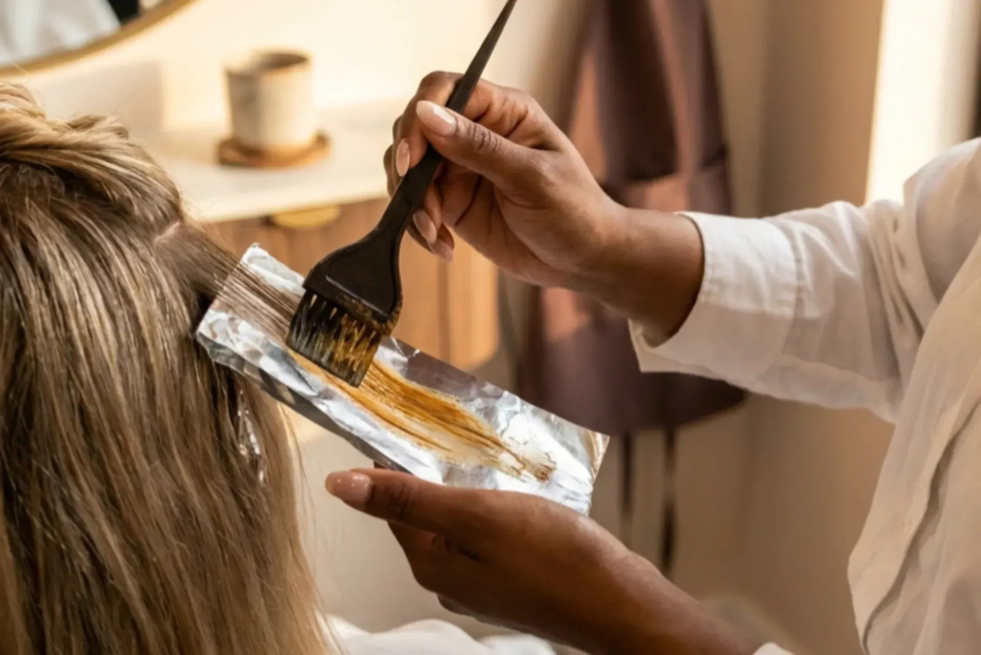 A colorist's hand painting color onto a foiled section of blonde hair with a tint brush in a warm salon