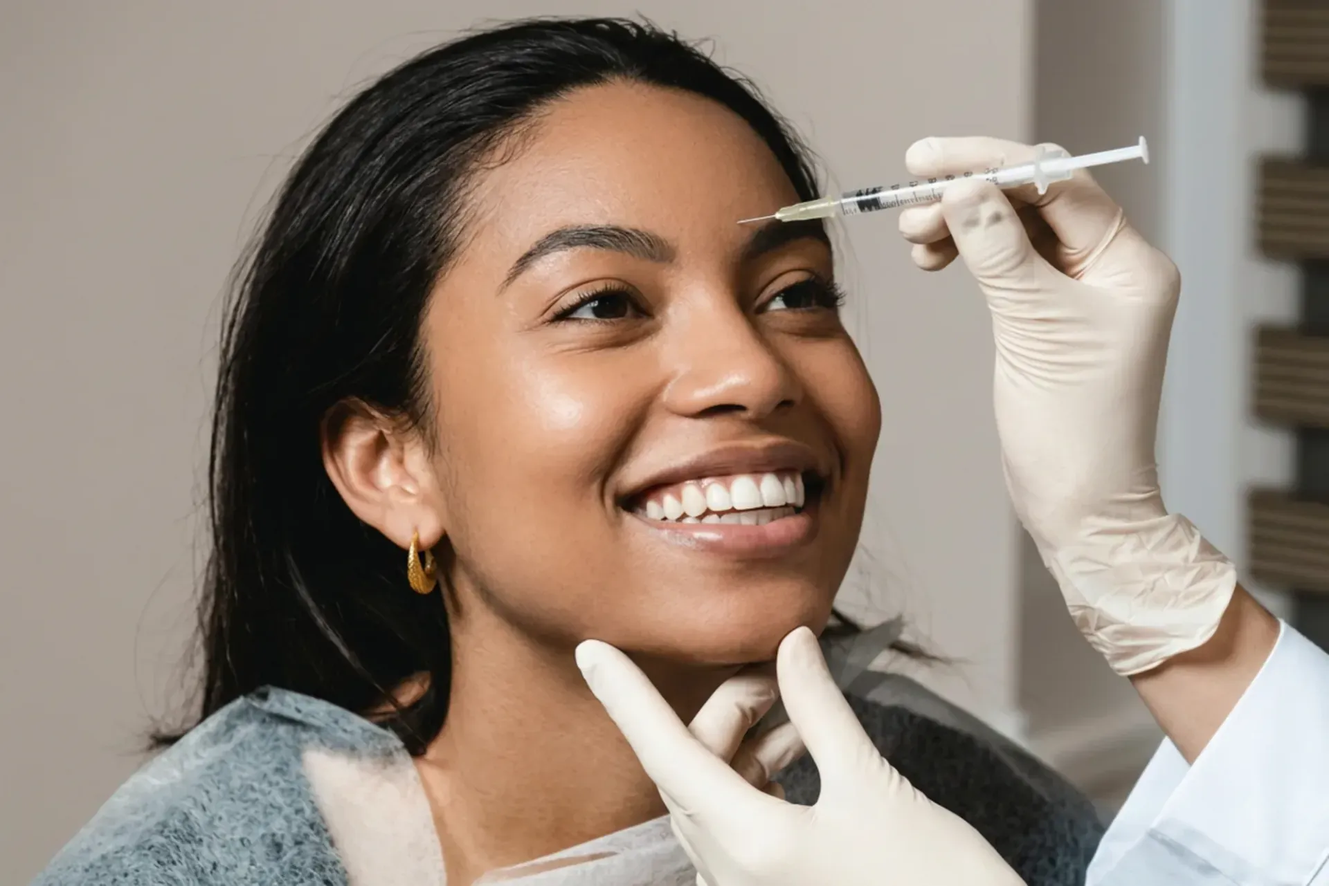 A smiling client receiving a cosmetic injection near her brow from a gloved clinician who steadies her chin