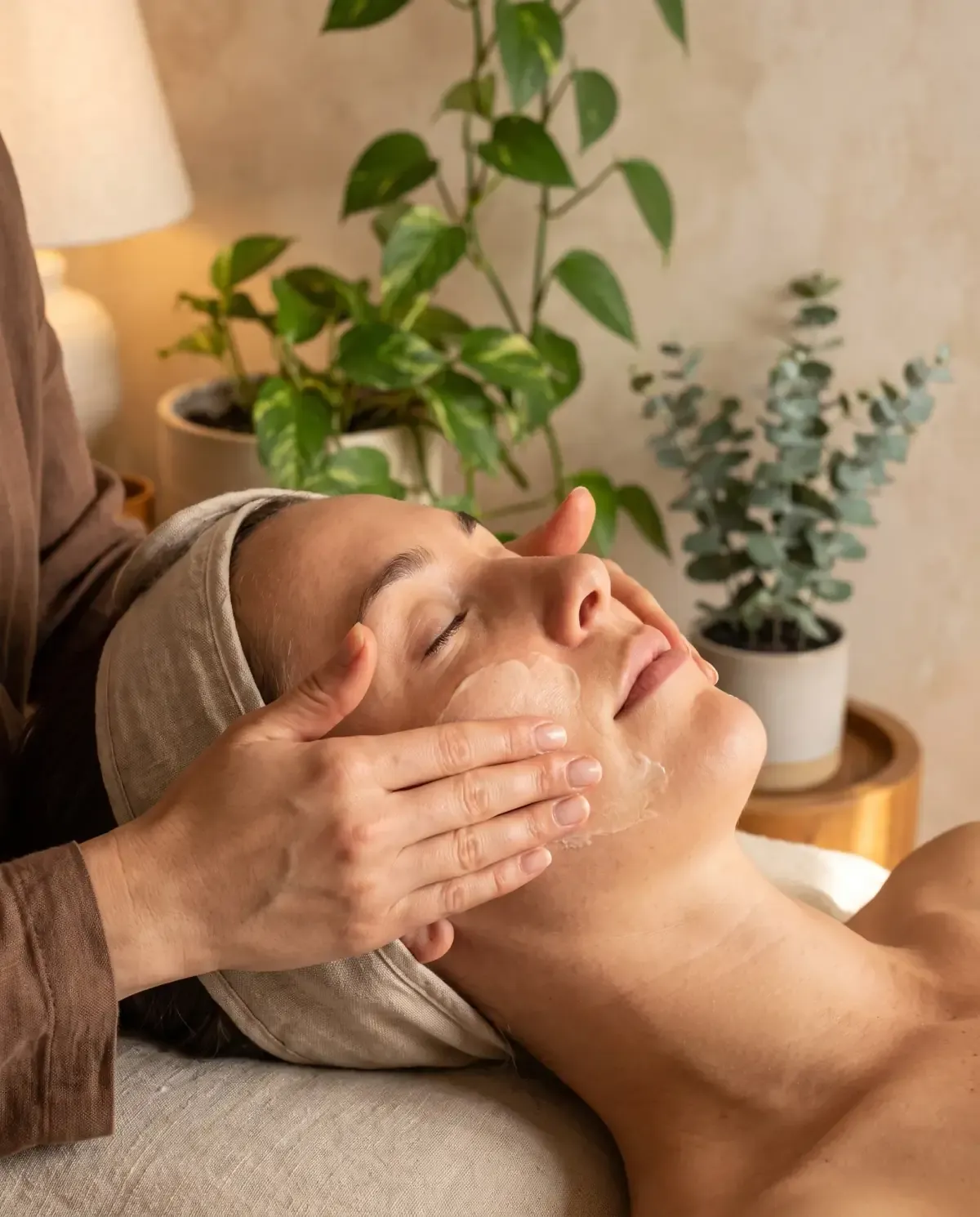 Warm colour mock photography: a close crop on a client receiving a facial, her eyes closed under a linen headwrap while fingertips work a cream along her cheekbone, a leafy pothos and a potted eucalyptus soft behind the bed